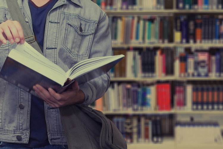 A student in a denim jacket holds open a textbook. The background is a shelf of books.