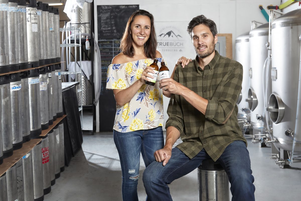 Louisa Ferrel CPA, CA and Conrad Ferrel CPA, CA pose in their workroom, each with a bottle of True Buch in their hands that they are clinking together.