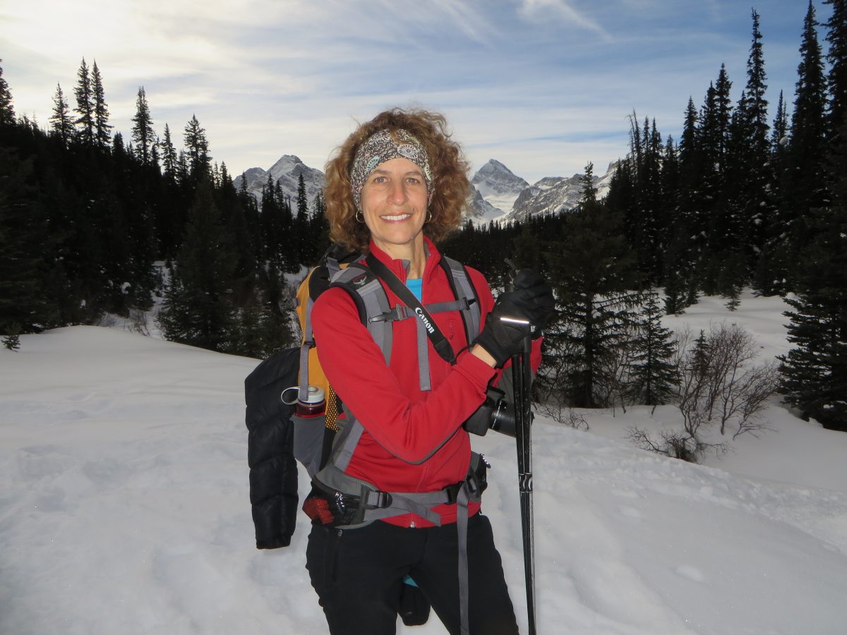 Viv Klingbeil CPA, CGA is wearing hiking gear and smiling at the camera. The landscape is snowy and there are trees and mountains behind her.