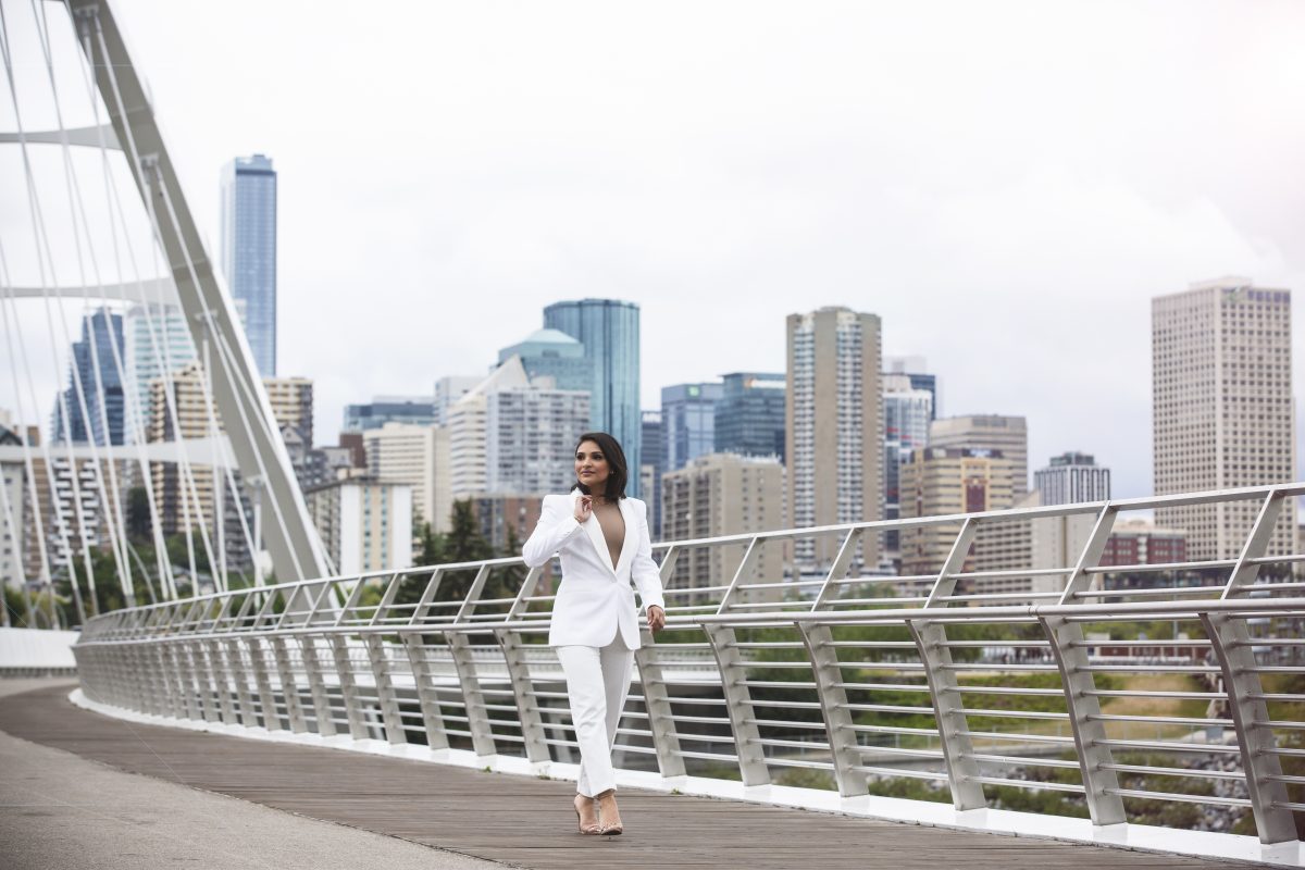 Priya Kapur CPA, CA is wearing a white fitted suit and walking along the Walterdale Bridge. The Edmonton skyline is behind her.