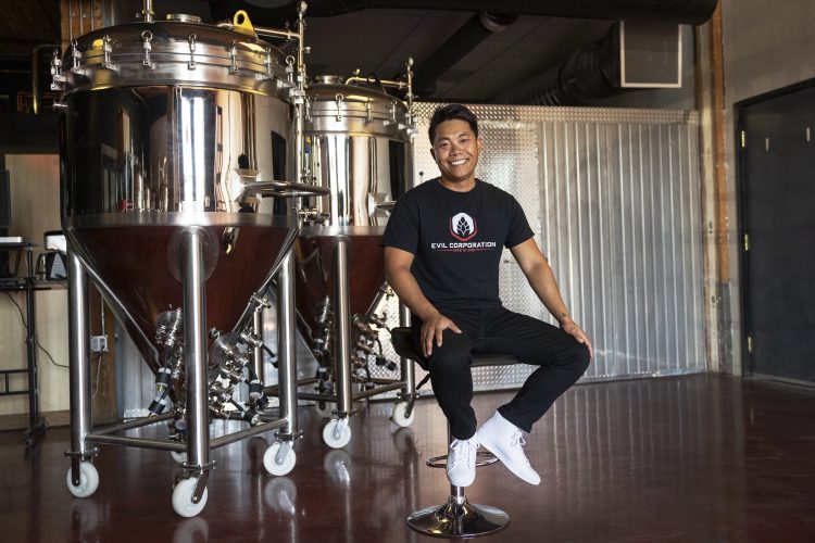 Quan Ly CPA, CA sits on a chair, smiling next to two fermenters.