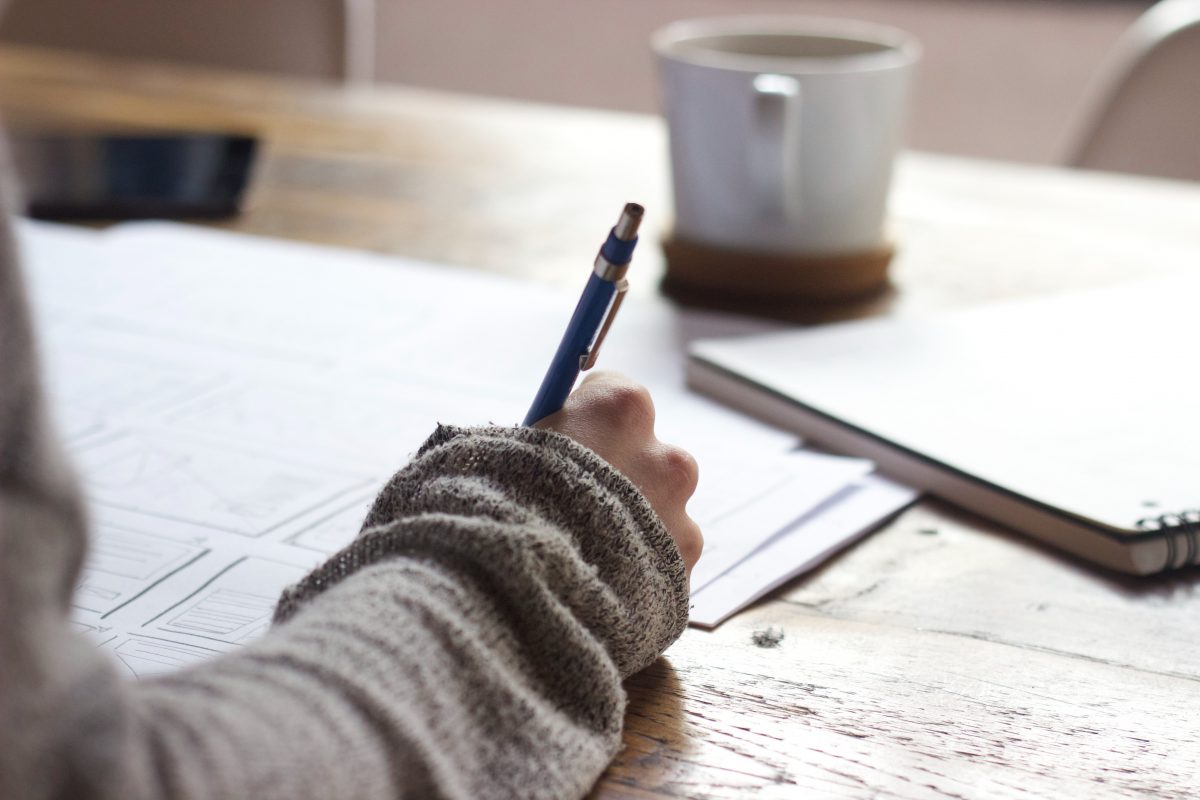 A hand is holding a pen and writing in a notebook. A cup of coffee is in the background.