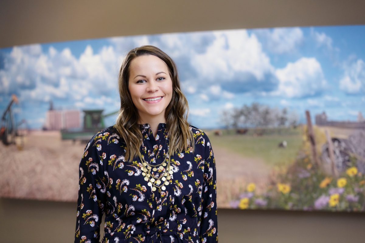 Kristy Jackson CPA, CA is wearing a blue floral blouse and smiling at the camera. Behind her is a mural of rural Alberta.