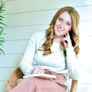 Jess Schultz rests her head on her hand and smiles at the camera. She is also writing in a notebook on her lap.