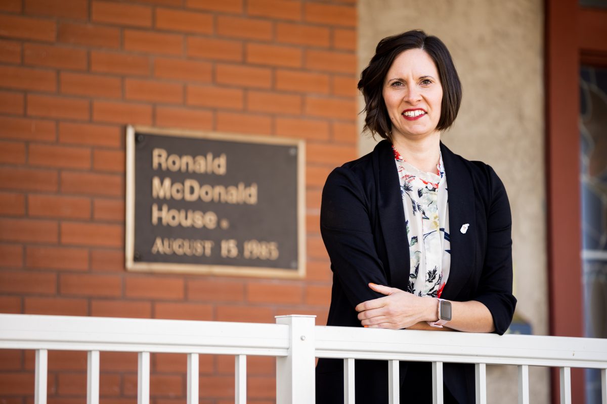 Desiree Bedard CPA, CGA stands in front of Ronald McDonald House. Her arms are crossed in front of her and leaning on a white railing.