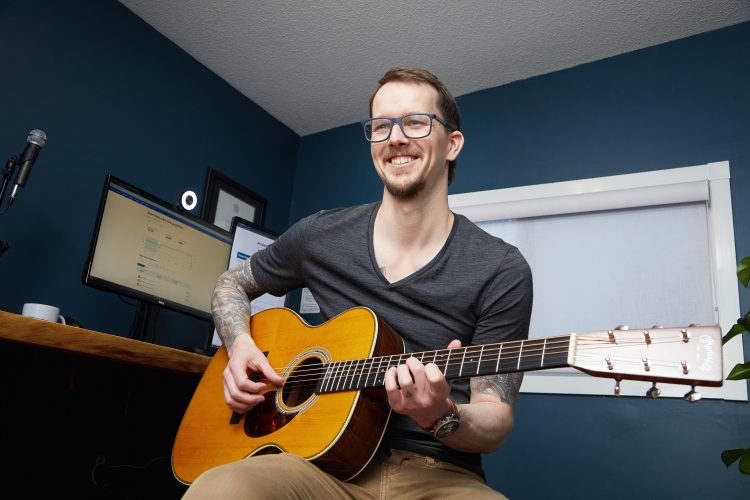 Joel Leclerc CPA, CGA smiles at the camera while playing a guitar. In the background is a computer with two monitors, a microphone and a webcam.