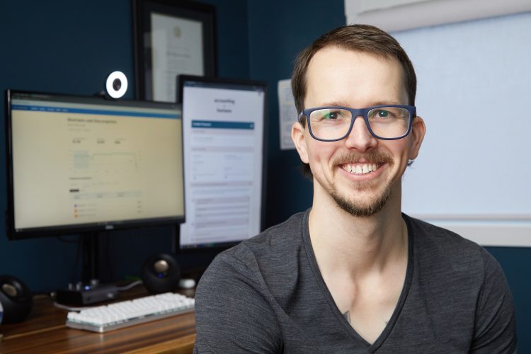 Joel Leclerc CPA, CGA smiles at the camera. In the background is a computer with two monitors and a webcam.