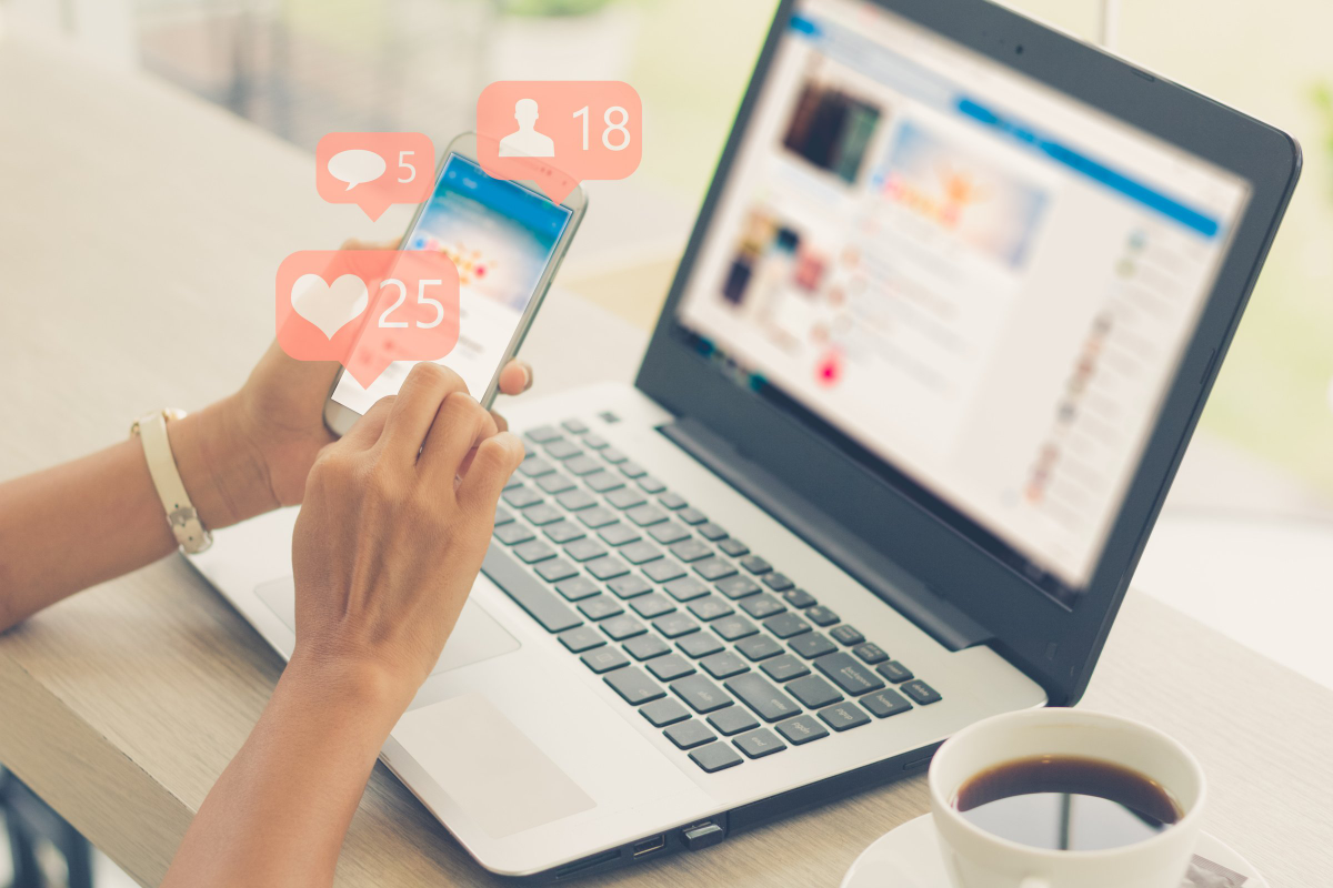 A person is on their phone on social media with a laptop and coffee cup in the background.