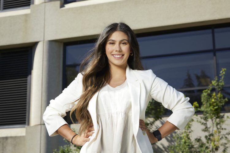 Emily Parker smiles at the camera with her hands on her hips. She is wearing white and a grey building with windows is behind her.
