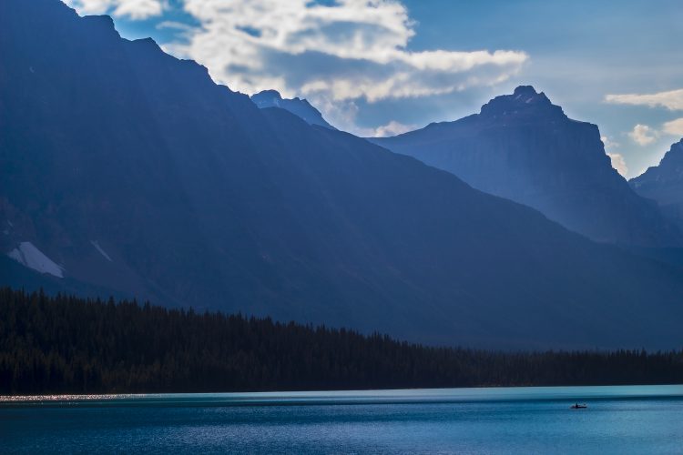 Mountains in the background with trees in the mid-ground and a lake in the foreground, all in shades of blue.