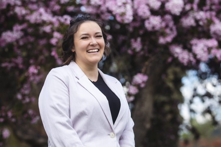 Sarah Deleon is smiling at the camera in front of a blooming tree. She is wearing a white blazer over a black shirt, and her short hair is wavy and half up.