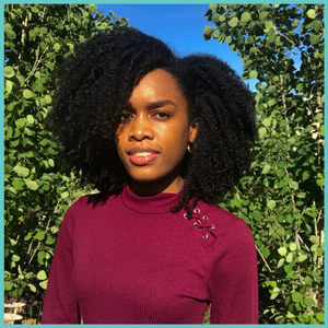 Jessica Grandison is smiling at the camera. Behind her is greenery. Her hair is natural and shoulder-length.