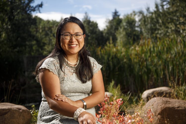 Cynthia Priest CPA, CMA smiles at the camera with her hands crossed in front of her. She is wearing a light grey lace dress and is outdoors.