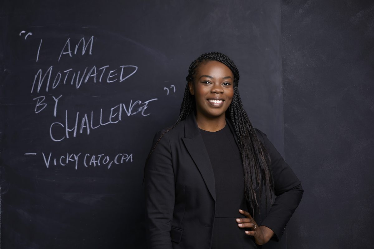 Vicky Cato CPA smiles at the camera. She is wearing all black, and has her hand on her hip. Behind her is a chalkboard with the text: I am motivated by challenge. - Vicky Cato CPA