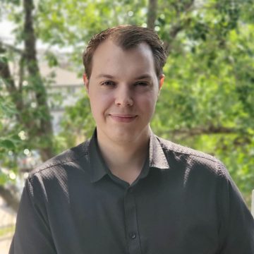 A person smiles at the camera. They have short brown hair and are wearing a black button-up shirt.
