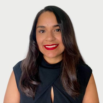 A person smiles at the camera. They are wearing red lipstick, have long dark hair, and are wearing a sleeveless black shirt.