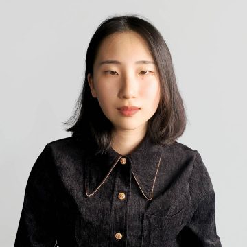 A person looks at the camera. They have shoulder-length dark hair and are wearing a dark button-up shirt with a large collar.