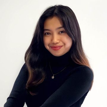 A person smiles at the camera. They have long dark hair and are wearing a black turtleneck.