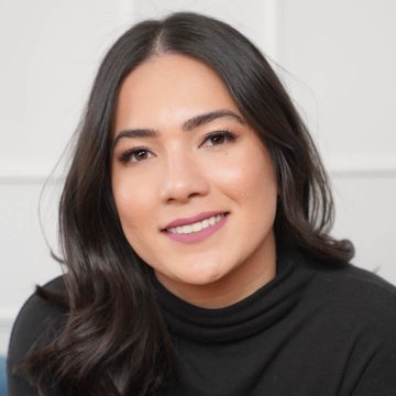 A person smiles at the camera. They have long dark hair and are wearing a black turtleneck.