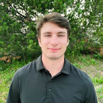 A person smiles at the camera. They have short brown hair and are wearing a black button-up shirt.