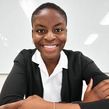 A person smiles at the camera with their arms crossed in front of them. They are wearing a white button-down shirt under an open black blazer.