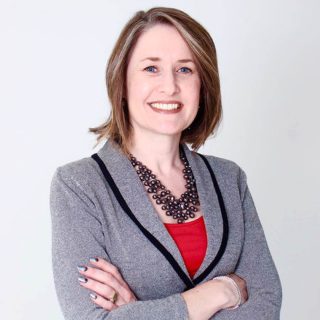 Kimberley Dart is smiling at the camera with her arms crossed in front of her. She is wearing a grey blazer over a red top.