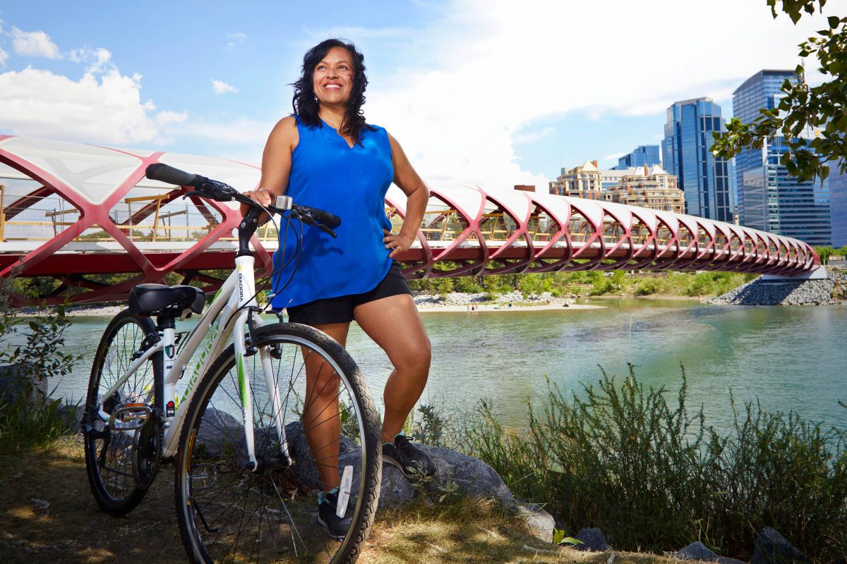 Shaheen Kassam CPA, CMA smiles off camera. She is posing with her bike next to a river with a bridge in the background.