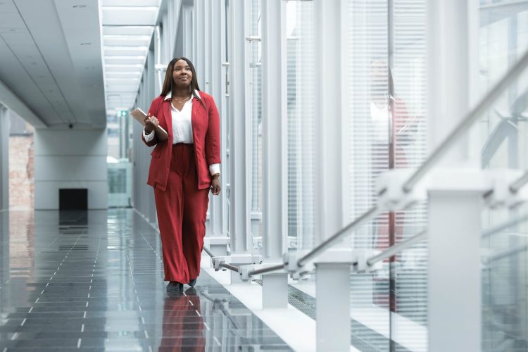 Kamilah Parker wearing a red business suit over a white button-down shirt. She has long dark hair and dark skin and is walking down a hallway with floor-to-ceiling windows.