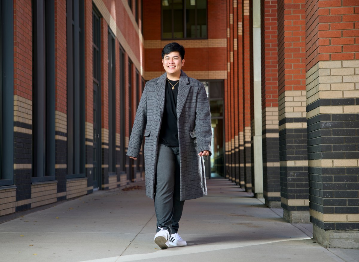 Al Kim Lutao is walking past a brick wall. He is wearing a long, grey jacket over a black top and is smiling.
