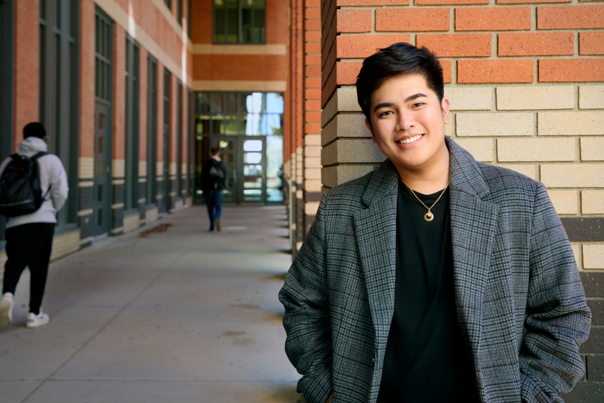 Al Kim Lutao is leaning against a brick wall. He is wearing a long, grey jacket over a black top and is smiling.