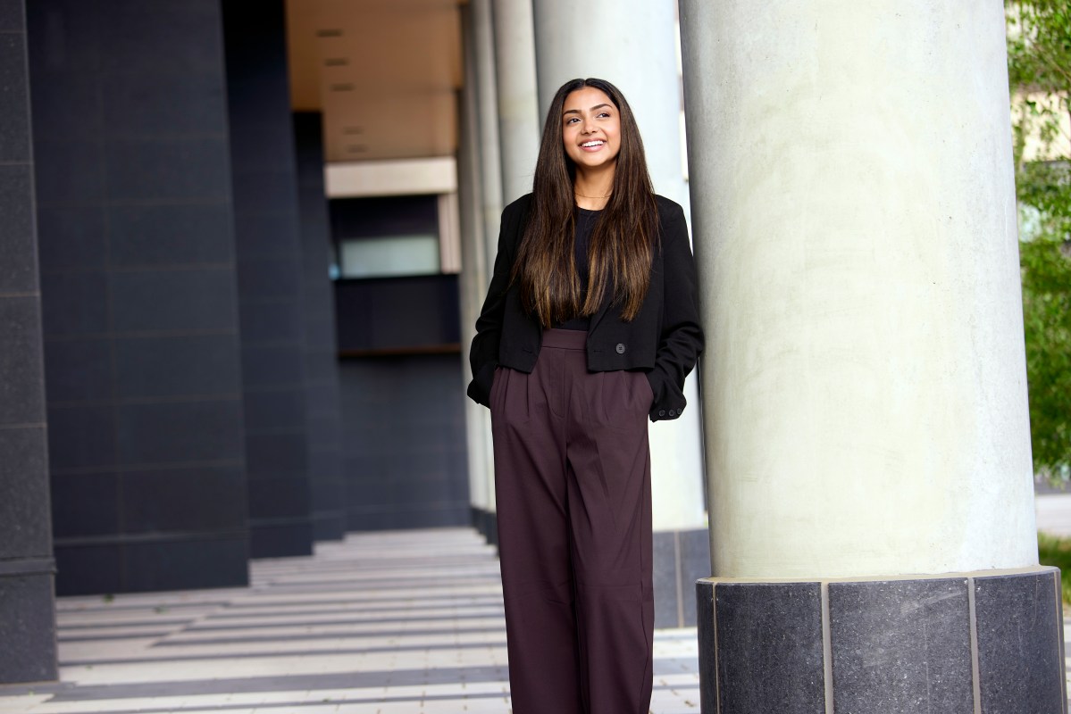 Gurleen Mangat poses leaning against a column with her hands in her pockets and a smile on her face. She is wearing dark purple pants and a black blazer.