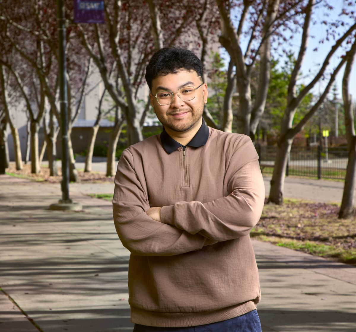 Marion Paul Mendoza poses in front of a row of trees with his arms crossed. He is wearing a brown sweater with a black collar showing from beneath it and glasses.